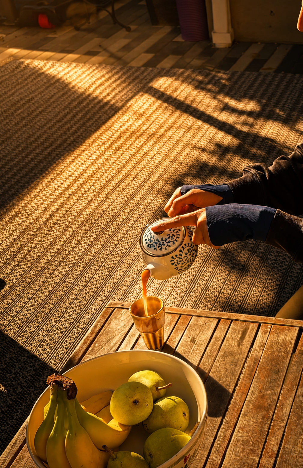 Hand pouring chai from a printed teapot into a brass cup in morning sunlight